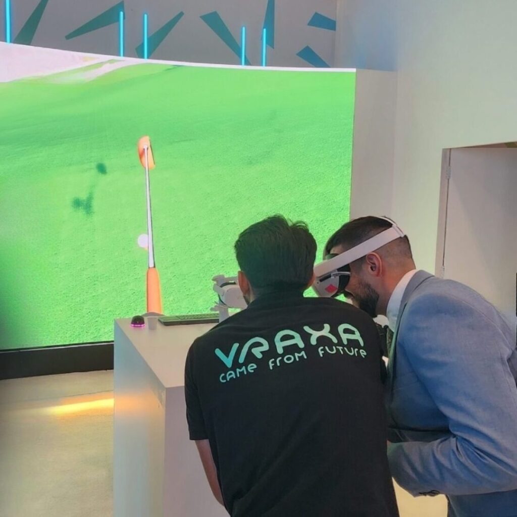 Three people engage with a virtual reality golf simulator. One person wears VR goggles, another observes, and a person with a VRAXA shirt is also focused on the display. The screen shows a golf course.