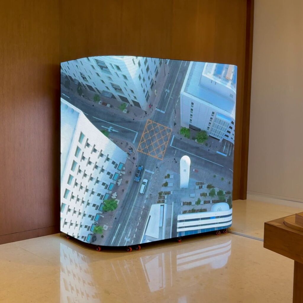 A 3D curved screen displays a high-resolution aerial view of a city intersection with buildings, roads, and vehicles. The screen is set against a wooden wall on a glossy tiled floor, creating a striking illusion.