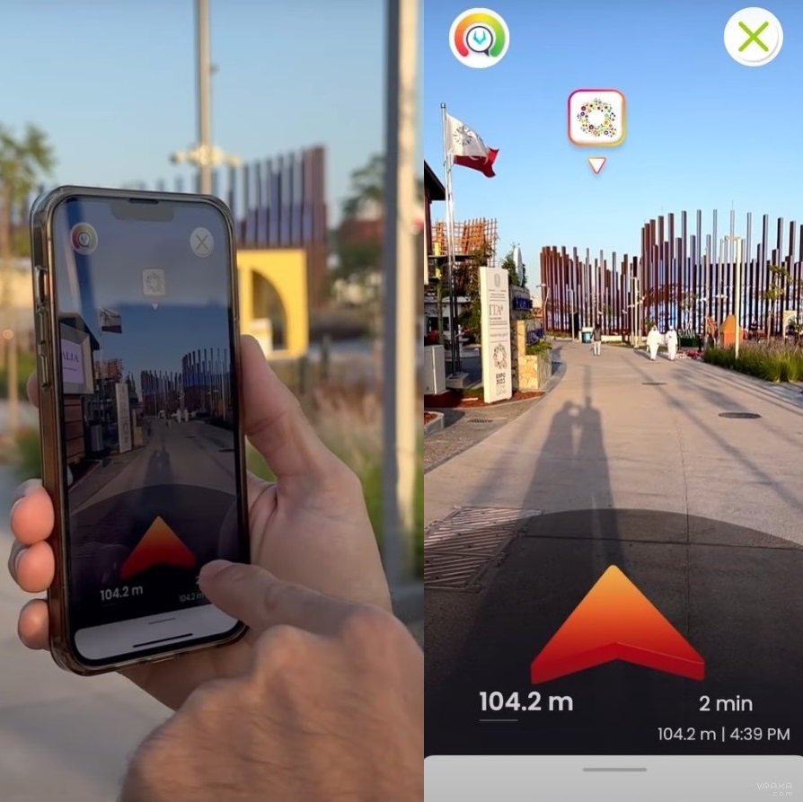 A person holds a smartphone displaying a navigation app with augmented reality, showing a path with a red arrow and distance. The real-world scene features a pathway lined with vertical poles and flags.