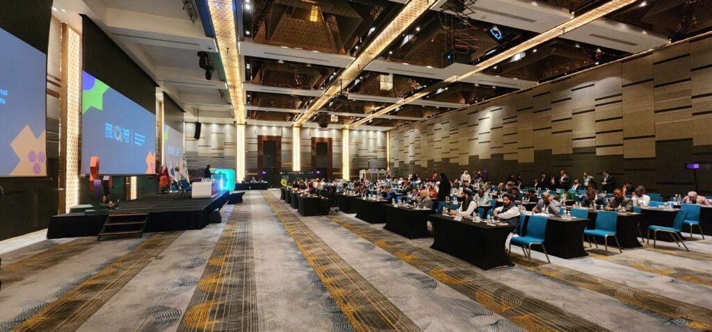 A large conference hall with many attendees seated at tables. Large screens display graphics and text at the front. The room has a high ceiling with ornate lighting and walls decorated with panels.