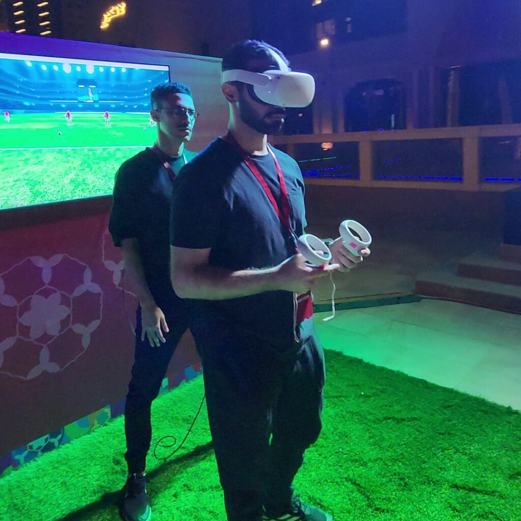 A person wearing a virtual reality headset and holding controllers stands on a grassy area indoors, with a large screen behind them casting the VR game. Another person stands nearby, watching.