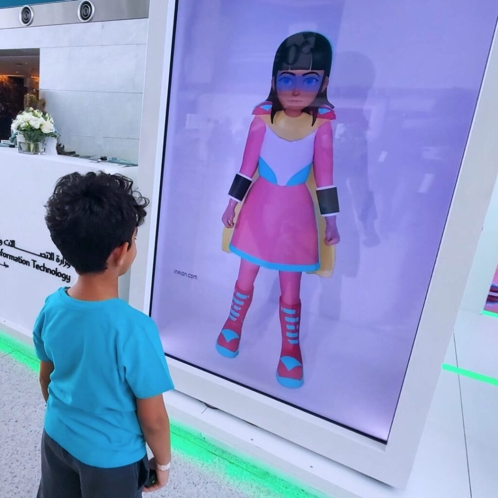 A child in a blue shirt stands in front of an interactive holobox displaying a cartoon character in a colorful outfit. The setting appears to be a technology exhibition with a booth and attendants nearby.