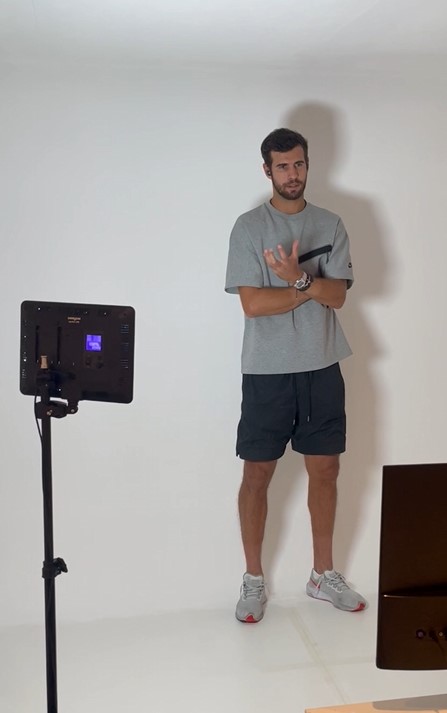 Man standing against a white wall in a studio. He is gesturing with one hand and looking towards a camera on a tripod in front of him. A light provider is positioned on the side.