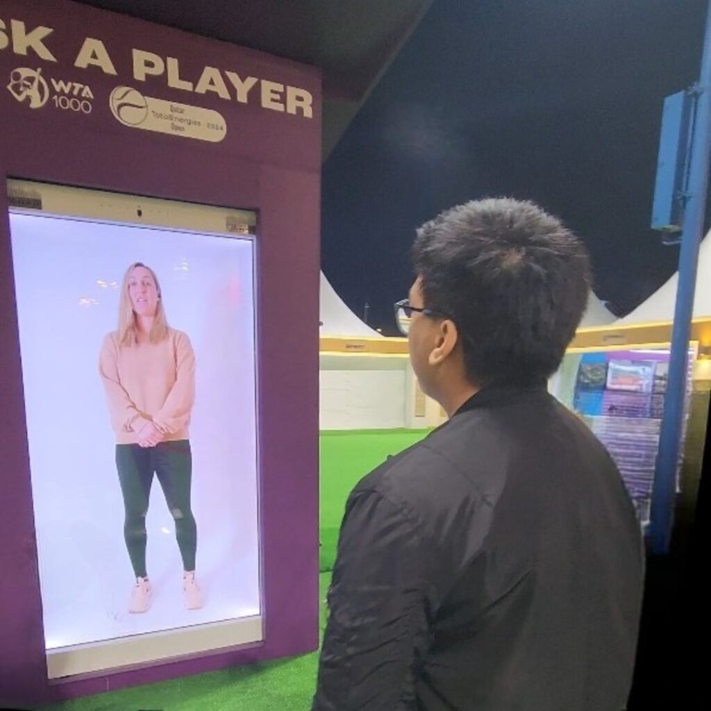 A person interacting with a large holobox featuring a virtually real woman in casual attire. The display is part of a tennis event. The setting is an outdoor venue at night.