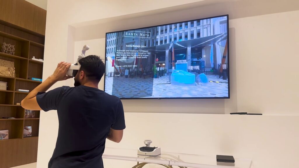 A person using a VR headset stands in front of a screen casting a virtual reality scene with a building and a text overlay. Shelves with books and a white brick wall are in the background.