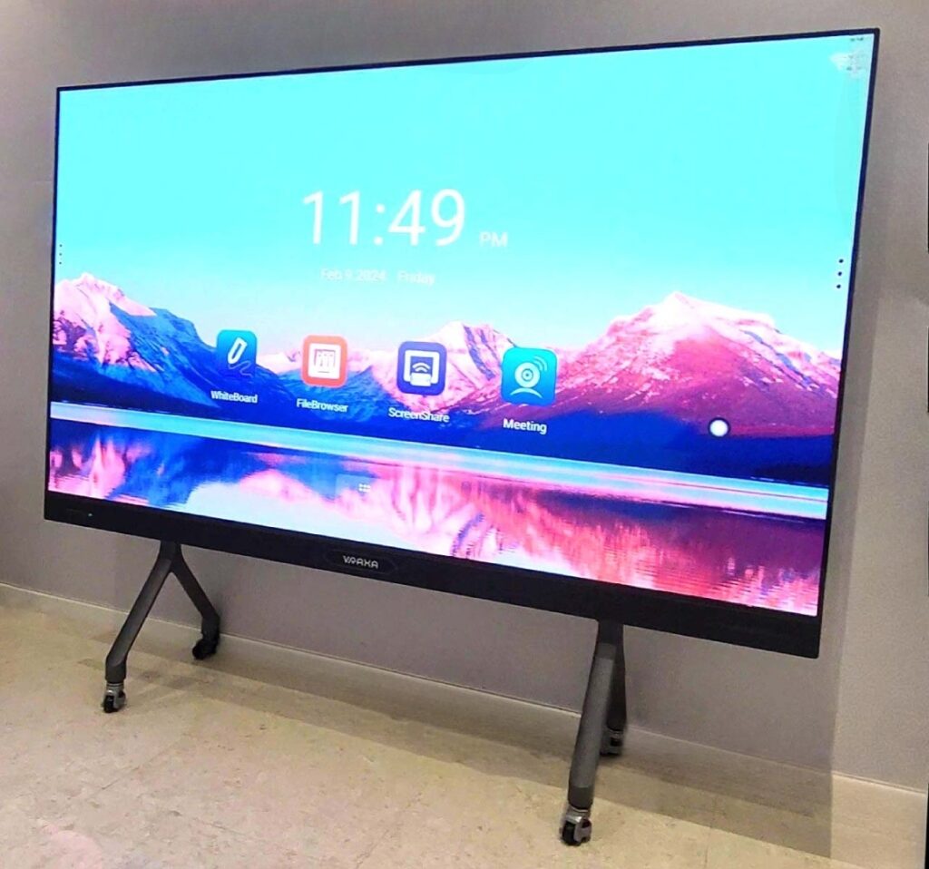 A large interactive touch screen on a stand displays a scenic wallpaper of mountains and a lake. The screen shows app icons for Whiteboard, File Browser, Screen Share, and Meeting.