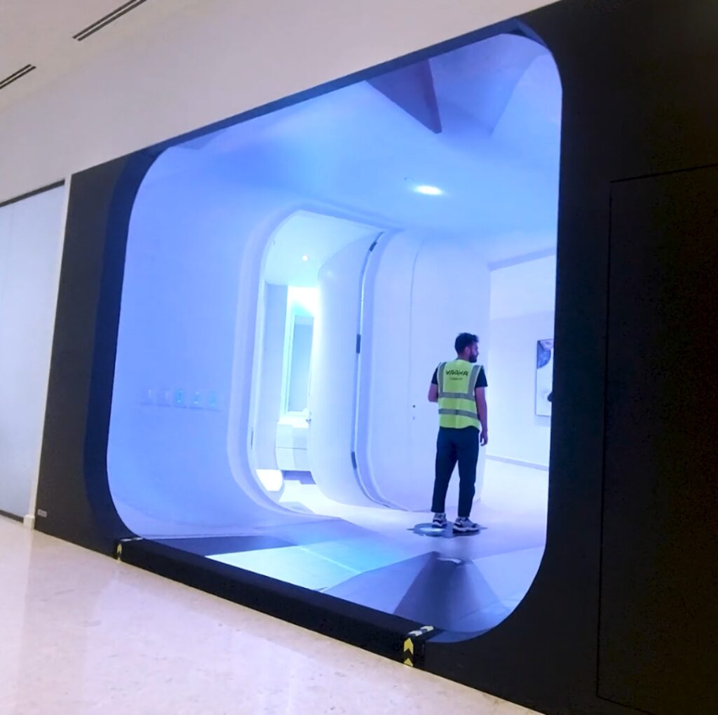 A man stands in a fully immersive room, showing a 360 degrees view of apartment. The surrounding area has a polished floor and white walls.