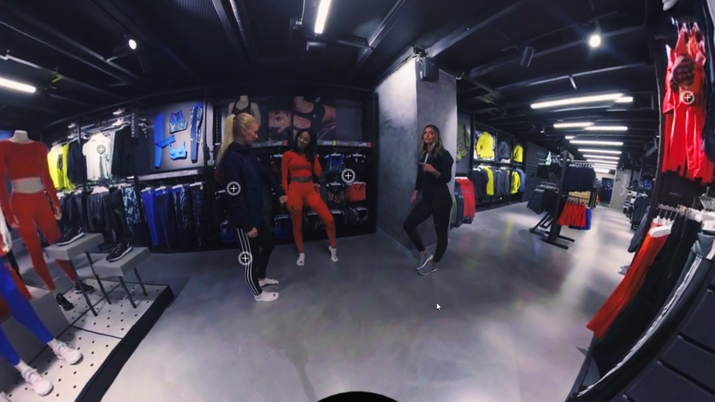 A 360-degree view of a sportswear store shows three people conversing. They are surrounded by racks of athletic clothing, including jackets and pants, in various colors. The store has bright lighting and modern decor.