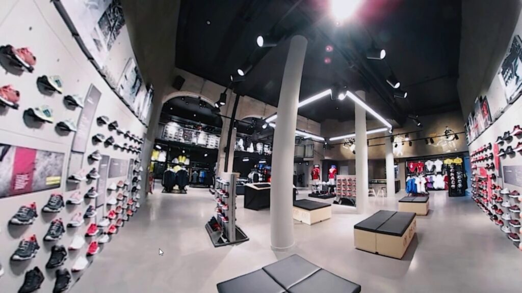 A 360-degree view of a modern shoe store with bright lighting and sleek design. Shoes are displayed on shelves lining the walls. The store features gray flooring, black seating, and two white pillars, with apparel visible in the background.