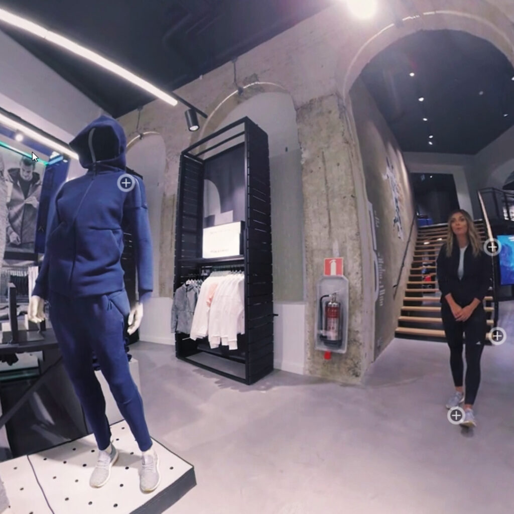 A 360-degree view of a clothing store interior with a mannequin wearing a blue tracksuit to the left and various garments on display. A person in casual attire stands to the right. The store has concrete walls and a staircase leading to another level.