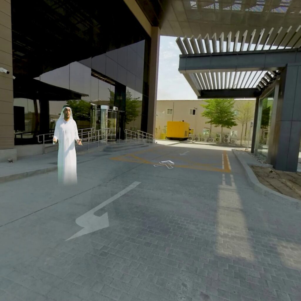 A preview of virtual experience showing a 3D avatar in a long white robe and headscarf is standing on a paved driveway near a modern glass building. Arrows are displayed on the ground, indicating virtual movements.