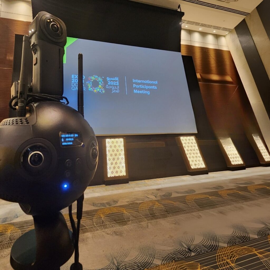 A 360-degree camera in the foreground faces a large screen displaying the Expo 2023 Doha Qatar logo and the text International Participants Meeting in a conference room.