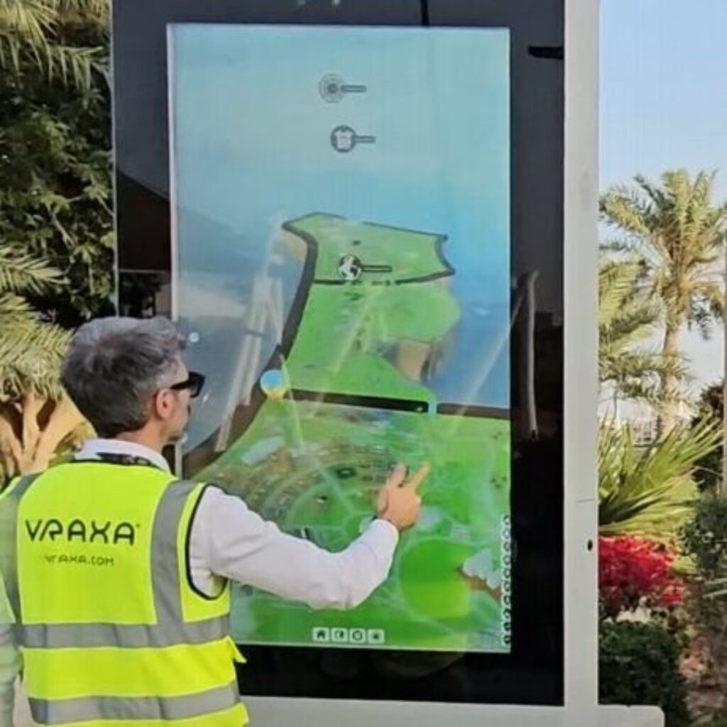 A person wearing a VRAXA vest interacts with a large interactive touchscreen outdoors. The screen displays a 3D model animation with bright colors.