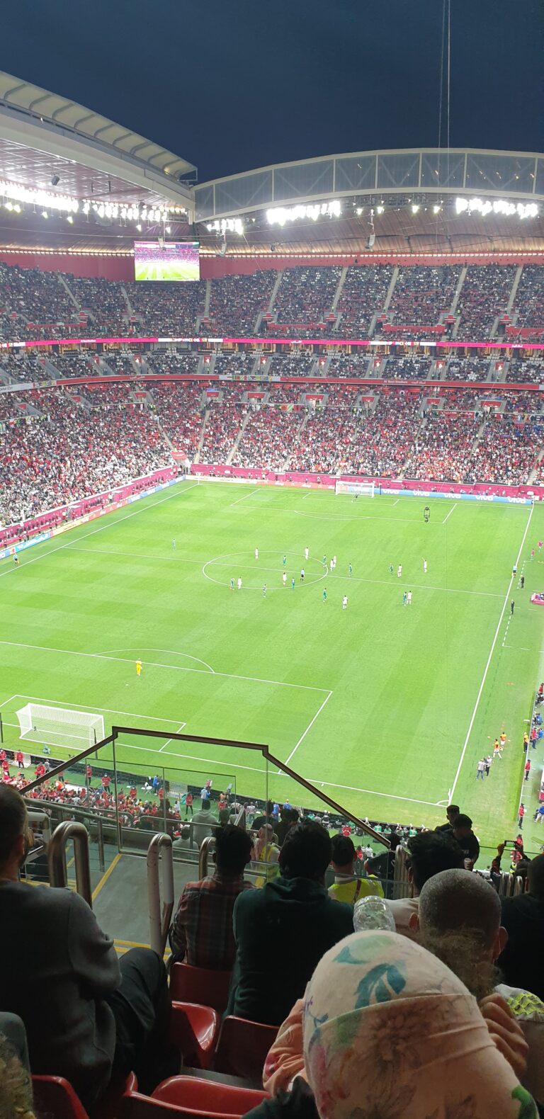 A packed football stadium at night. Fans fill the stands as two teams play on a brightly lit green field. Stadium lights are on, and spectators are visible in the foreground, wearing various colors and watching the match.