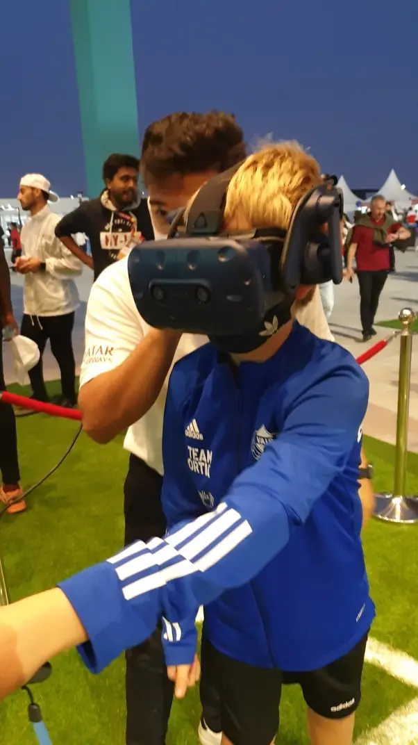 A child wearing a virtual reality headset and holding controllers is guided by another person on a grassy area. The guide is wearing a white shirt with Qatar Airways on it. They are indoors with onlookers in the background.