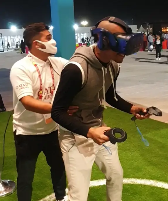 A person wearing a virtual reality headset and holding controllers is guided by another person on a grassy area. The guide is wearing a white shirt with Qatar Airways on it. They are indoors with onlookers in the background.