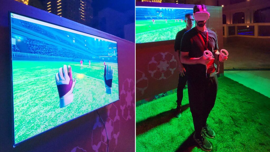 A person wearing a VR headset and holding controllers stands on artificial grass. A screen displays a virtual soccer field, showing gloved hands in view, simulating goalkeeping. Another person stands in the background.