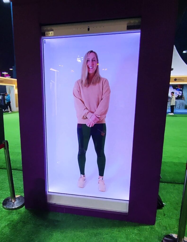 A woman stands virtually inside a large Holobox from Vraxa at an outdoor event, wearing a pink sweater, black pants, and pink shoes. The screen emits a soft purple light against the green flooring.