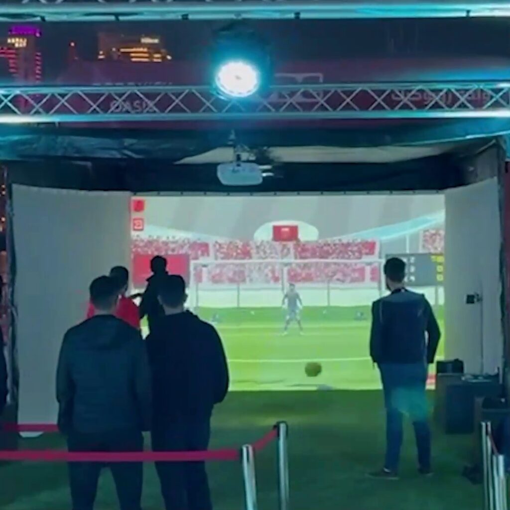 People are playing interactive soccer game projected on a 3-side projection screens. The scene shows a soccer field with goalposts and a goalkeeper on the screen. A small group of players stands nearby, with one taking a shot.