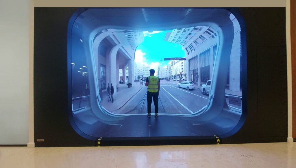 A person stands in front of a large screen displaying a street scene with buildings and a tram. The screen creates an immersive effect, resembling a window to the outside city. It is a fully immersive room, 4 walls, ceiling and floor.