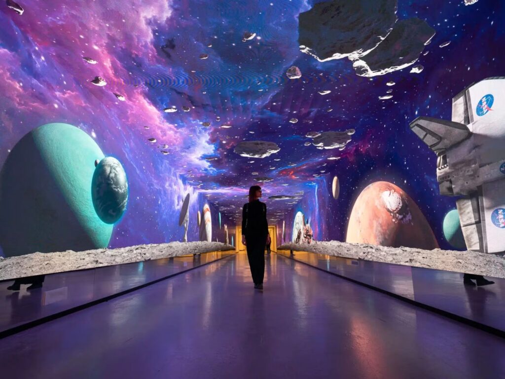 A person walks down a futuristic hallway with a vibrant, cosmic display on the ceiling and walls. The scene includes colorful planets and asteroids in a galaxy setting, creating an immersive space experience akin to those found in cutting-edge setups across Qatar, UAE, and KSA.