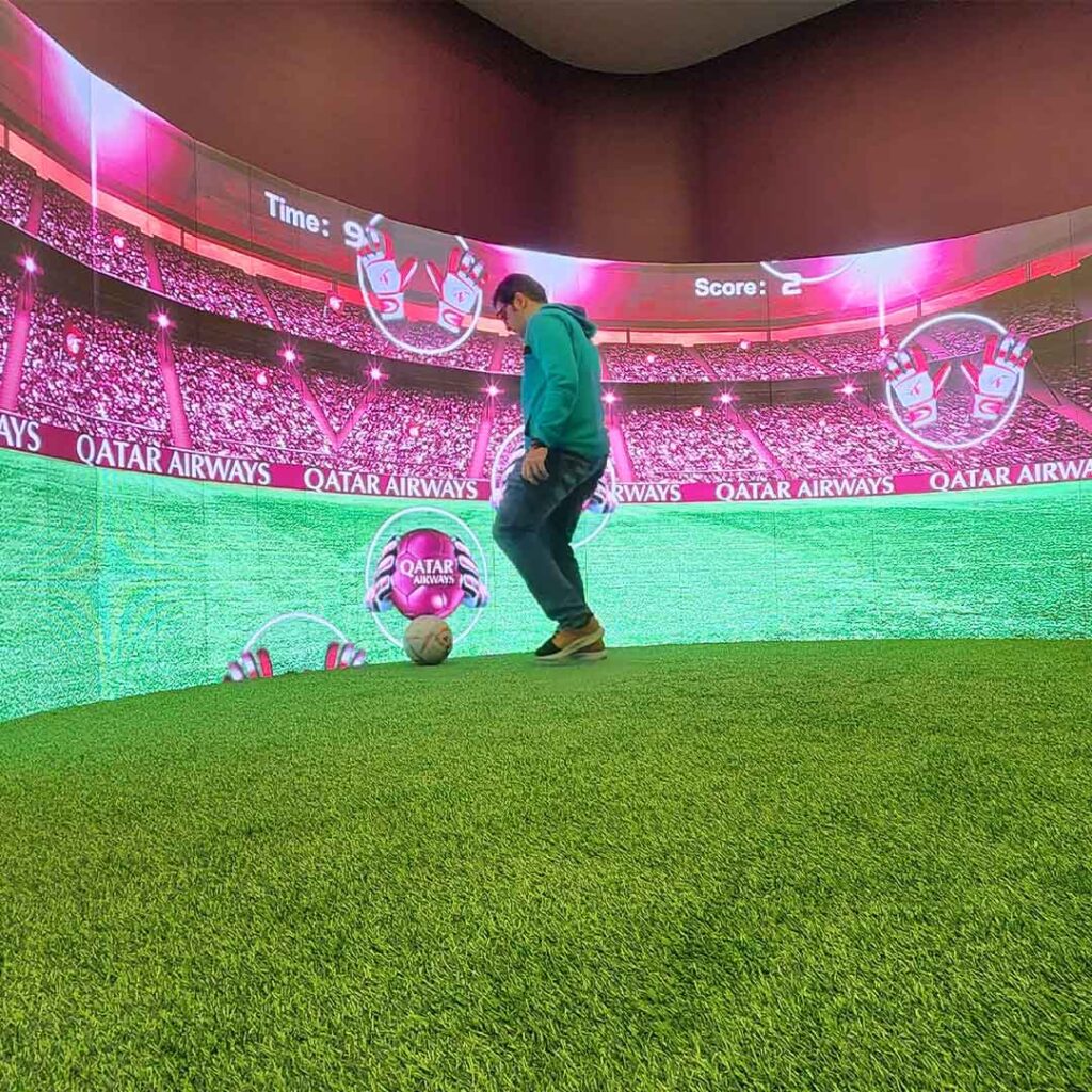 Person in a green hoodie plays soccer on a small artificial turf field indoors, with a large interactive curved screen displaying a stadium background, scoreboard showing Time: 90, Score: 2, and Qatar Airways logo.