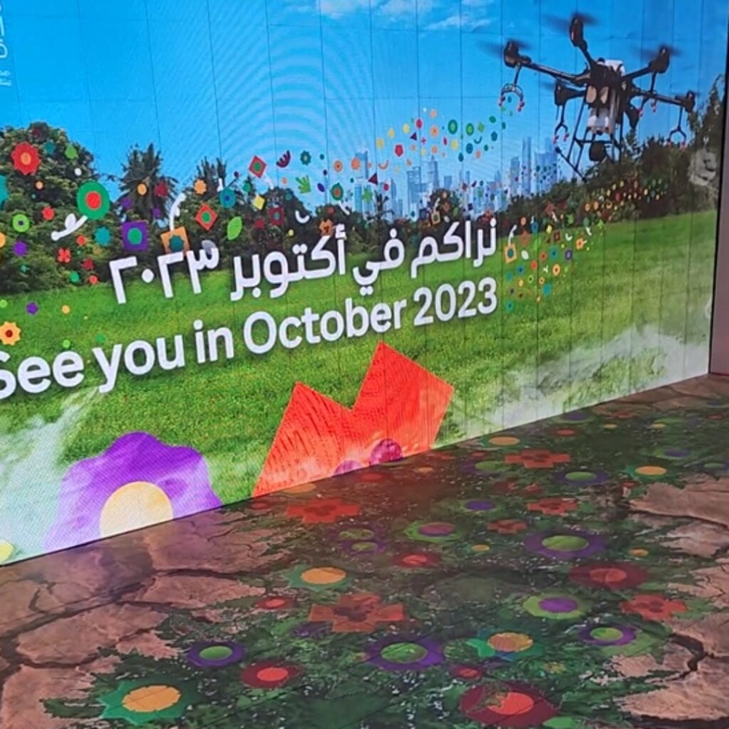 A digital interactive floor and wall display shows colorful flowers, a drone, and a city skyline with the text See you in October 2023.