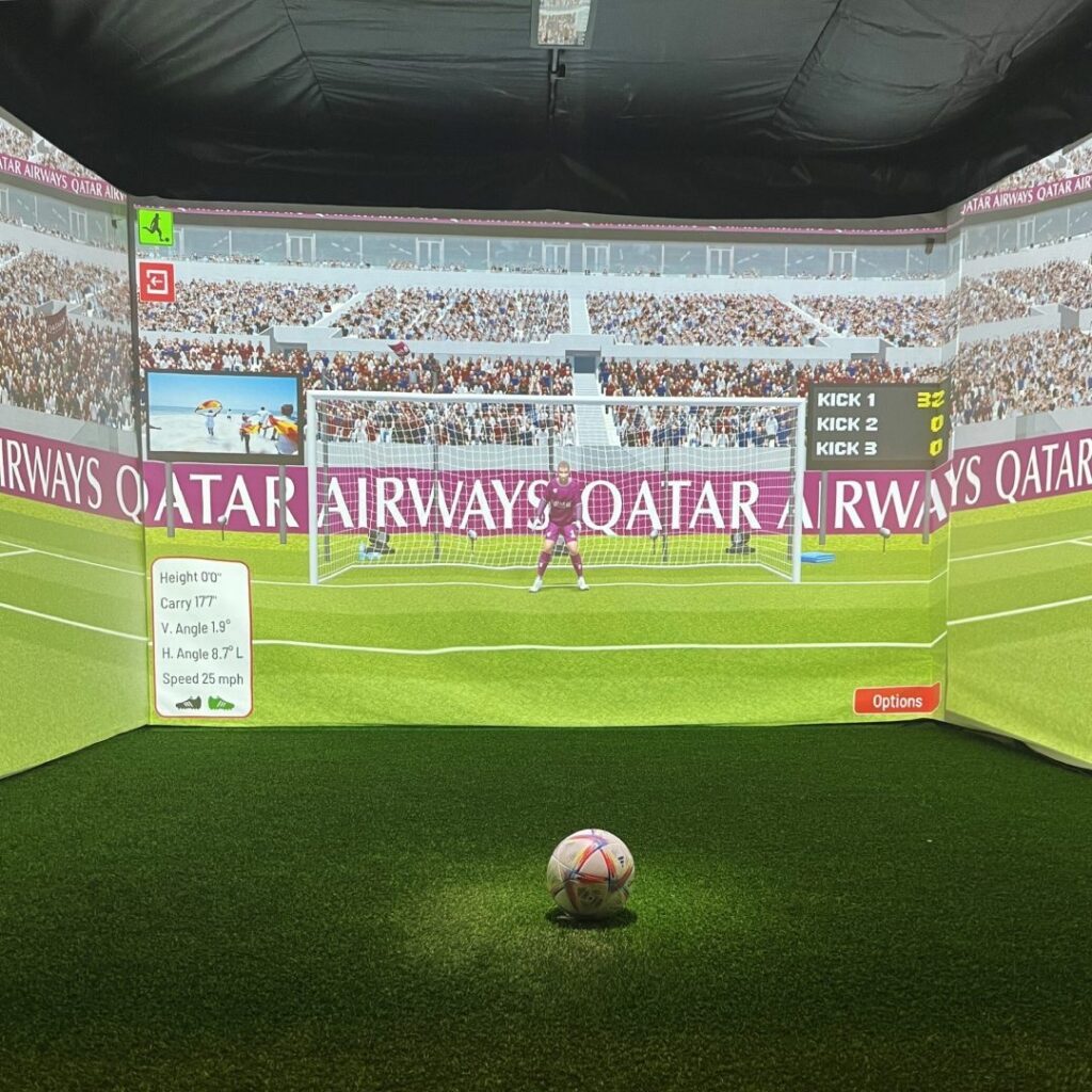 A football simulation setup using 3-side projection shows a digital goalkeeper and goal on a screen. A real football ball lies on artificial turf in the foreground. On-screen stats include ball height, angle, and speed. The backdrop displays a stadium with Qatar Airways advertisements.