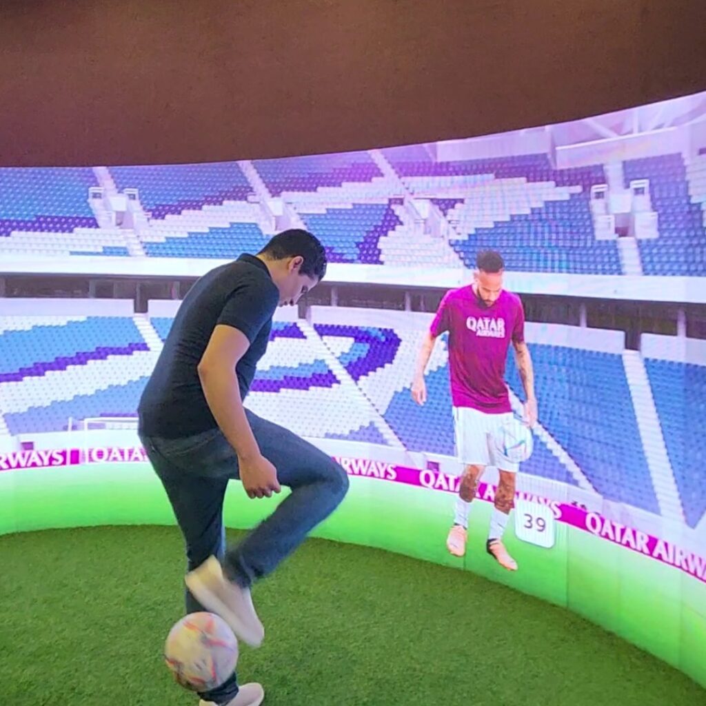 A man balances a football on his foot in front of a large interactive curved screen. The screen displays a football stadium with a player interacting with the man.