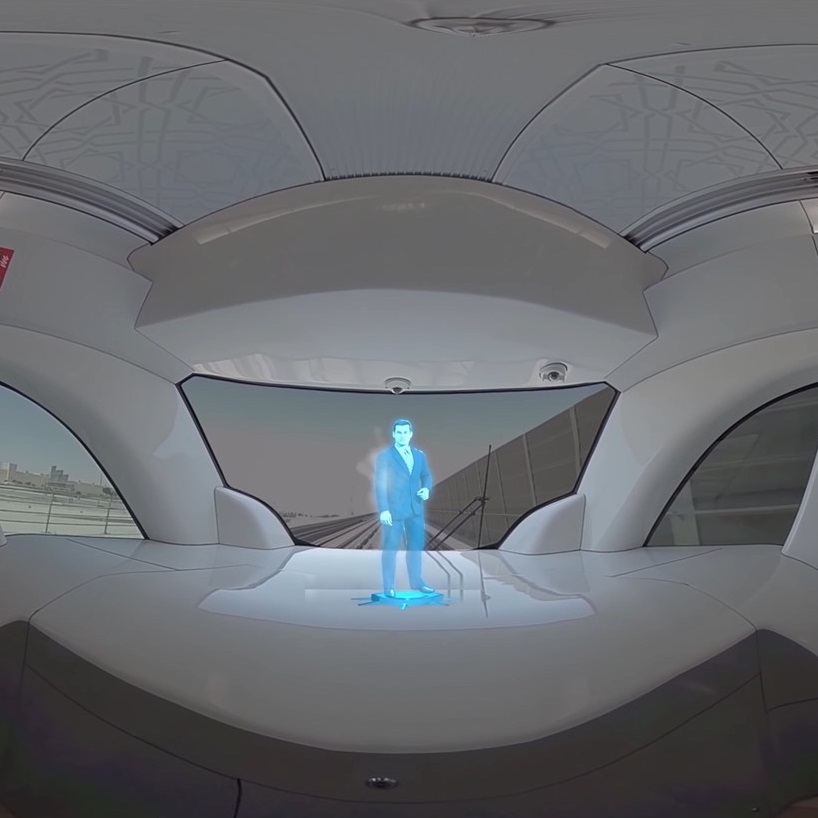 A futuristic train interior with sleek white design features a small blue hologram of a person standing near the front window, which shows a blurred cityscape view outside.