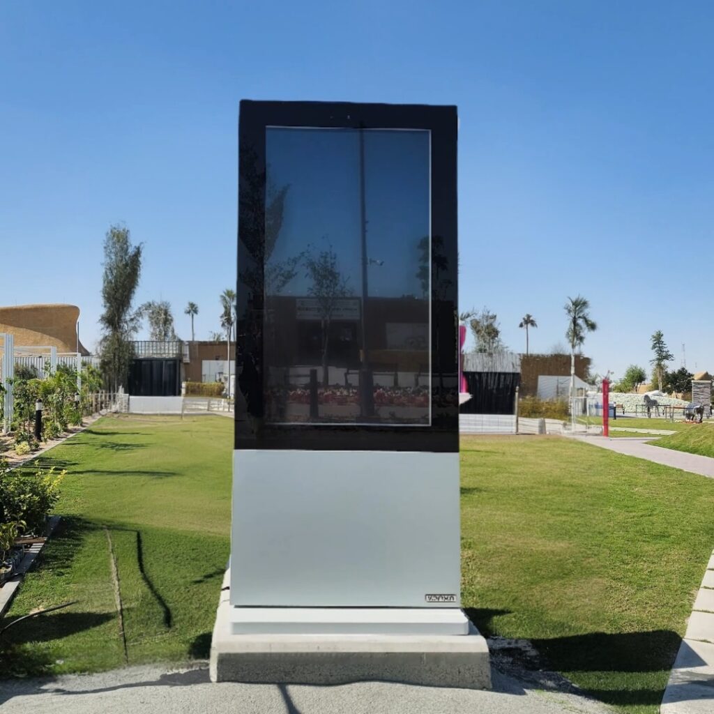 A large, vertical digital display stands on a concrete base in an outdoor park setting. The screen is turned off, reflecting the sky and surrounding trees. The area is sunny, with green grass and a clear blue sky in the background.