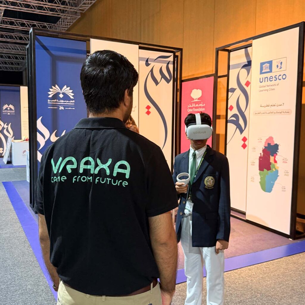 A person wearing a black VRAXA shirt observes another person using a VR headset at an exhibition. The display panels in the background feature intricate designs and the UNESCO logo.