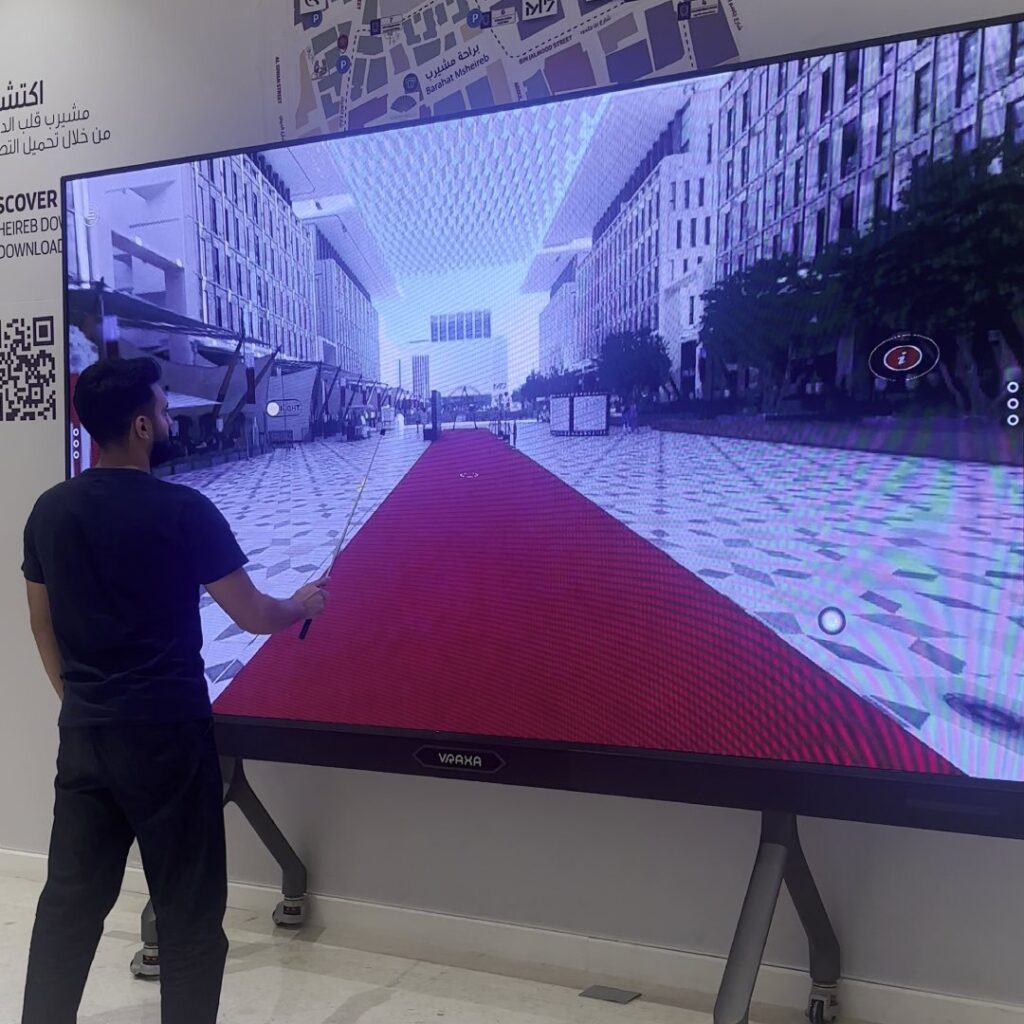 A person is interacting with a large interactive touch screen display that shows a virtual cityscape with a red carpet. They hold a long pointer stick, engaging with the content. The surrounding environment appears to be a modern indoor space.