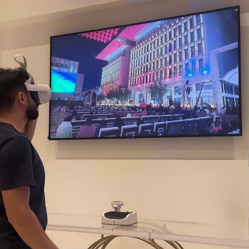 A person wearing virtual reality headset stands in front of a large wall-mounted TV casting an image of a building lit with colorful lights and a large audience seated outdoors. A gaming console is on a table below the TV.