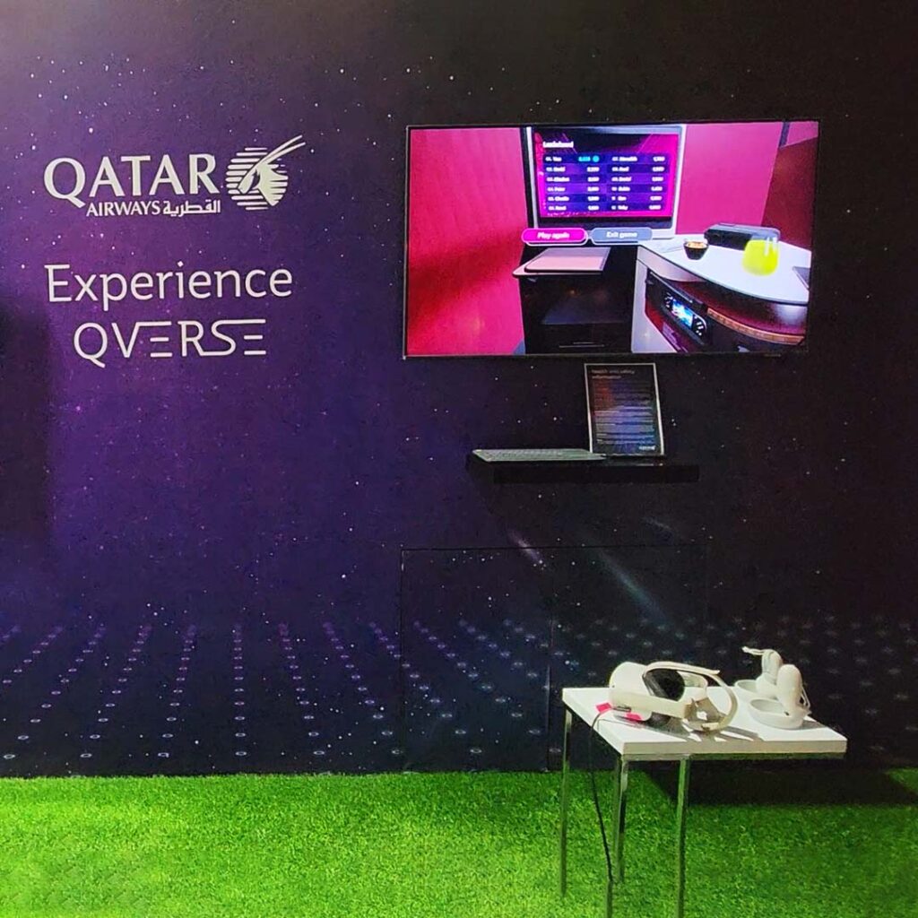 A Qatar Airways booth displays a screen and virtual reality headset on a small table on a grassy floor. The wall reads Experience Qverse with the airline logo. The screen shows a cabin interior, indicating a virtual experience.