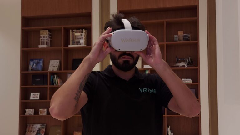 A person wearing a black shirt with a logo is immersed in Vraxa services using a VR headset labeled VRAXA in a cozy room adorned with wooden shelves brimming with books and decorative items.