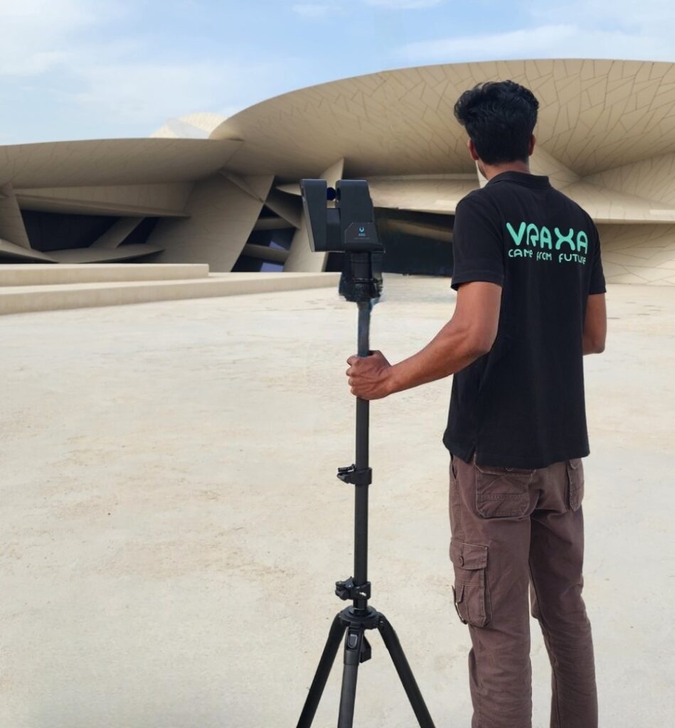 A person in a black T-shirt with green text stands outside a modern, abstract building about Vraxa Qatar, operating a camera on a tripod. Beside them is a suitcase. The sky is clear, highlighting the buildings curved, overlapping structures.