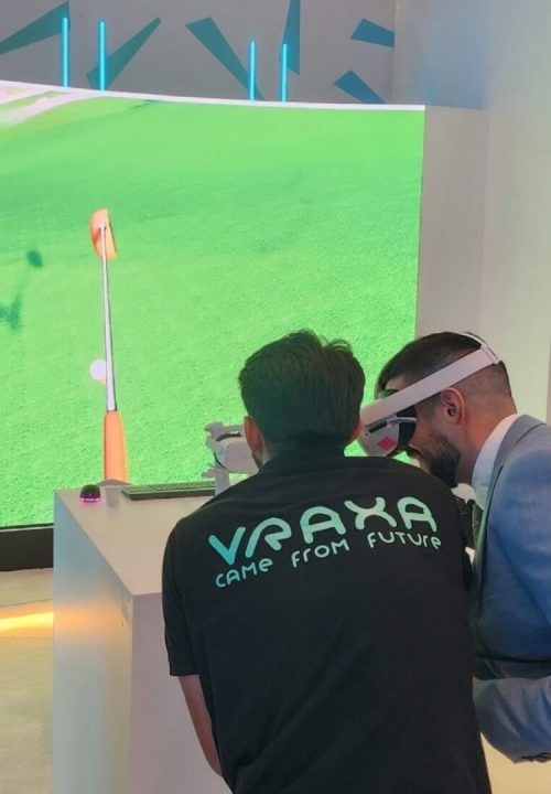 Three people engage with a virtual reality golf simulator. One person wears VR goggles, another observes, and a person with a VRAXA shirt is also focused on the display. The screen shows a golf course.