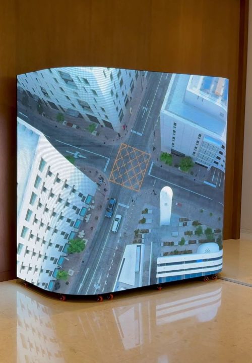 A 3D curved screen displays a high-resolution aerial view of a city intersection with buildings, roads, and vehicles. The screen is set against a wooden wall on a glossy tiled floor, creating a striking illusion.