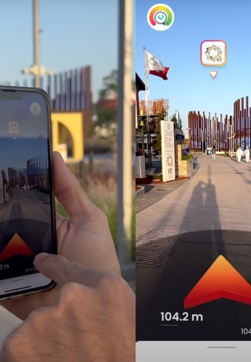 A person holds a smartphone displaying a navigation app with augmented reality, showing a path with a red arrow and distance. The real-world scene features a pathway lined with vertical poles and flags.