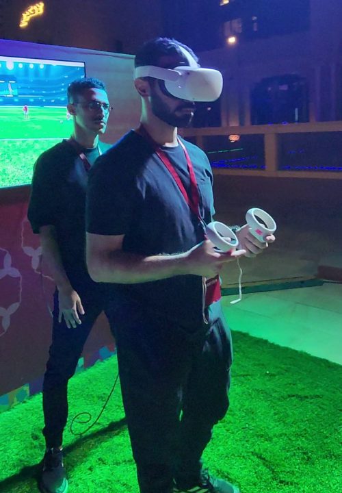 A person wearing a virtual reality headset and holding controllers stands on a grassy area indoors, with a large screen behind them casting the VR game. Another person stands nearby, watching.