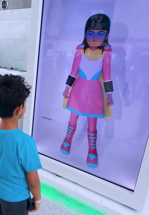 A child in a blue shirt stands in front of an interactive holobox displaying a cartoon character in a colorful outfit. The setting appears to be a technology exhibition with a booth and attendants nearby.