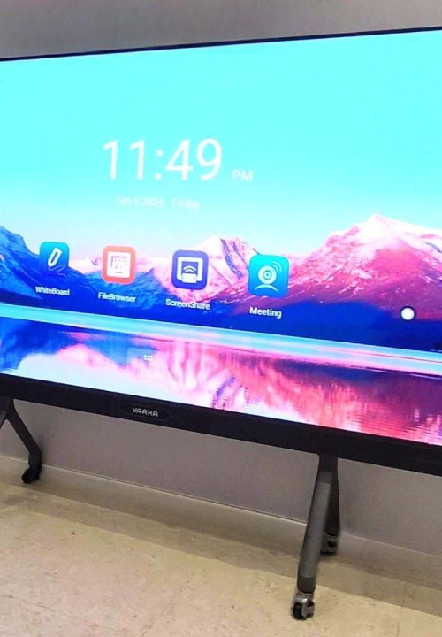 A large interactive touch screen on a stand displays a scenic wallpaper of mountains and a lake. The screen shows app icons for Whiteboard, File Browser, Screen Share, and Meeting.