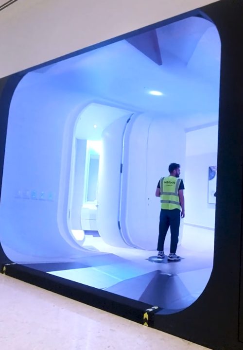 A man stands in a fully immersive room, showing a 360 degrees view of apartment. The surrounding area has a polished floor and white walls.