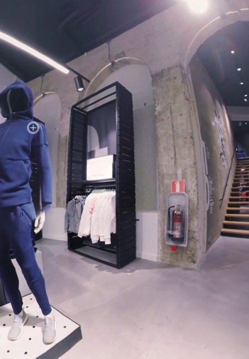 A 360-degree view of a clothing store interior with a mannequin wearing a blue tracksuit to the left and various garments on display. A person in casual attire stands to the right. The store has concrete walls and a staircase leading to another level.