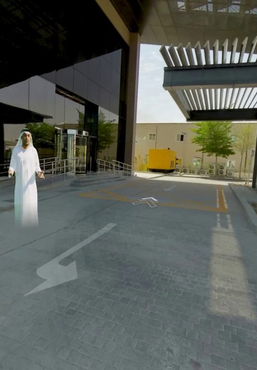 A preview of virtual experience showing a 3D avatar in a long white robe and headscarf is standing on a paved driveway near a modern glass building. Arrows are displayed on the ground, indicating virtual movements.