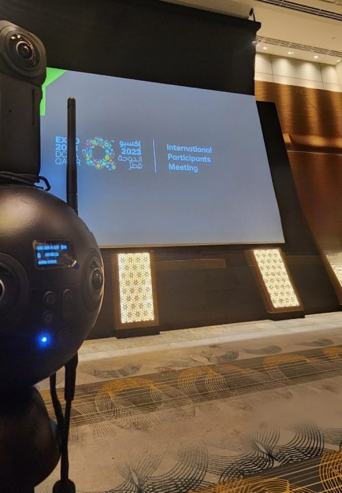 A 360-degree camera in the foreground faces a large screen displaying the Expo 2023 Doha Qatar logo and the text International Participants Meeting in a conference room.