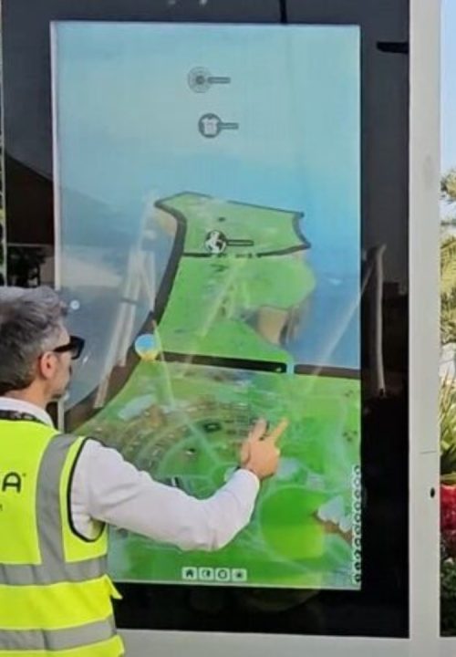 A person wearing a VRAXA vest interacts with a large interactive touchscreen outdoors. The screen displays a 3D model animation with bright colors.