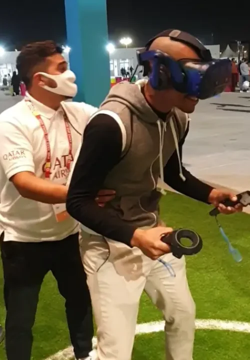 A person wearing a virtual reality headset and holding controllers is guided by another person on a grassy area. The guide is wearing a white shirt with Qatar Airways on it. They are indoors with onlookers in the background.