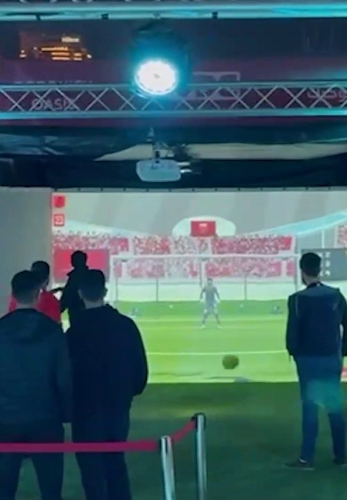 People are playing interactive soccer game projected on a 3-side projection screens. The scene shows a soccer field with goalposts and a goalkeeper on the screen. A small group of players stands nearby, with one taking a shot.